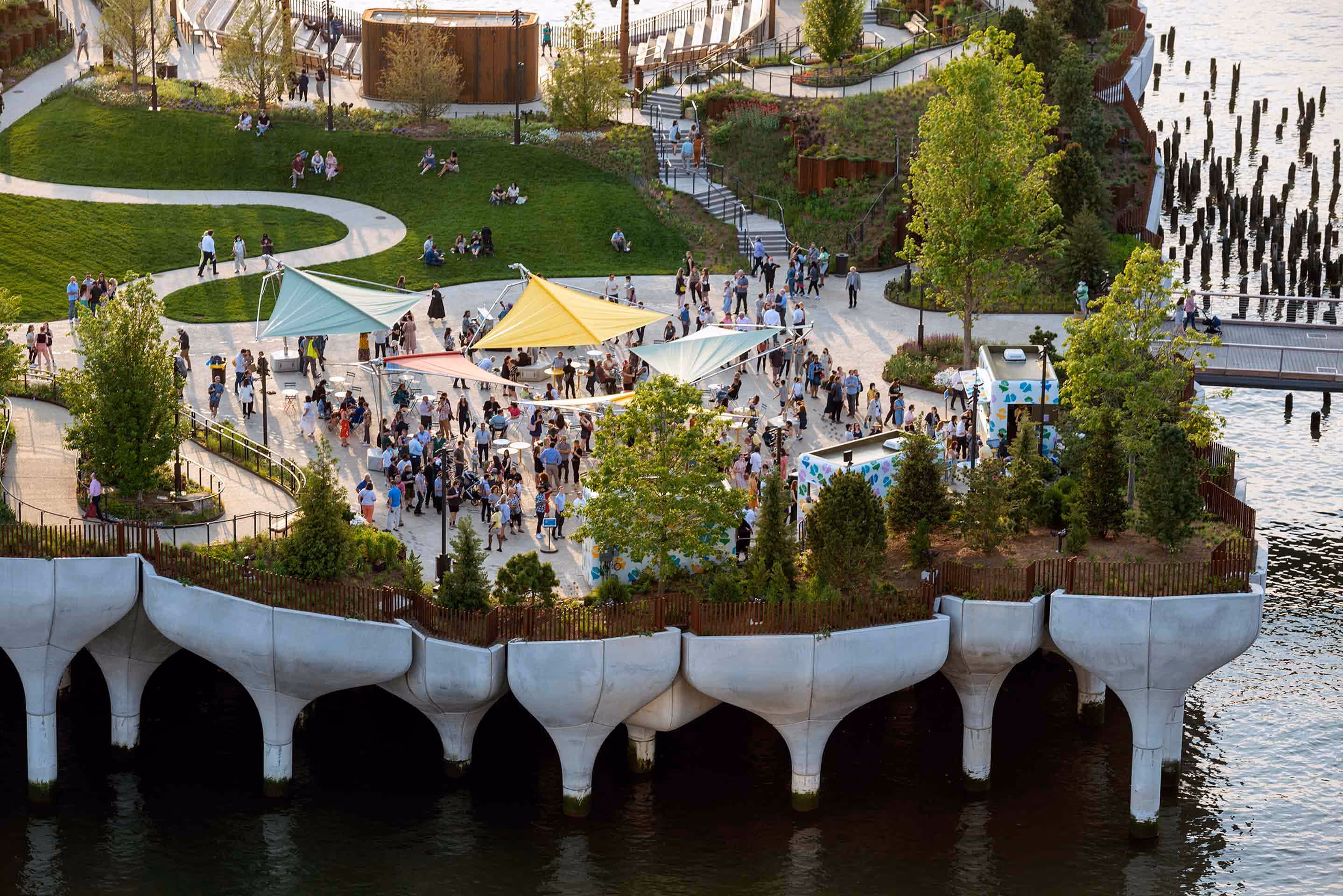 Aerial view of Little Island in New York with SunSquare Fold & Roll sailing umbrellas above a crowd surrounded by green vegetation, curving paths, and distinctive concrete pillars in the water.