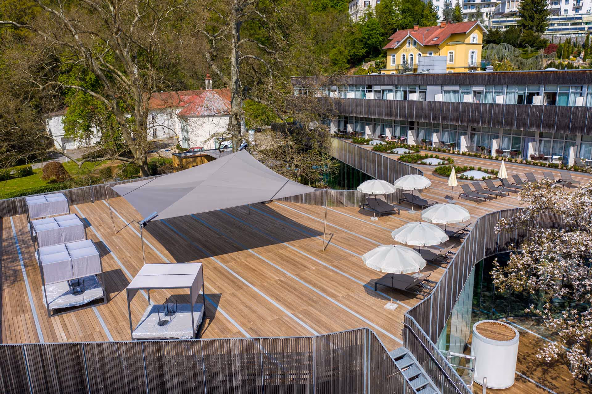 Luftaufnahme einer modernen Hotelterrasse mit großzügigem Holzdeck, zahlreichen Liegestühlen und SunSquare Sonnensegel. Ein großes, dreieckiges SunSquare-Sonnensegel spendet Schatten, während ein angrenzender Pool und stilvolle Glaswände den exklusiven Out