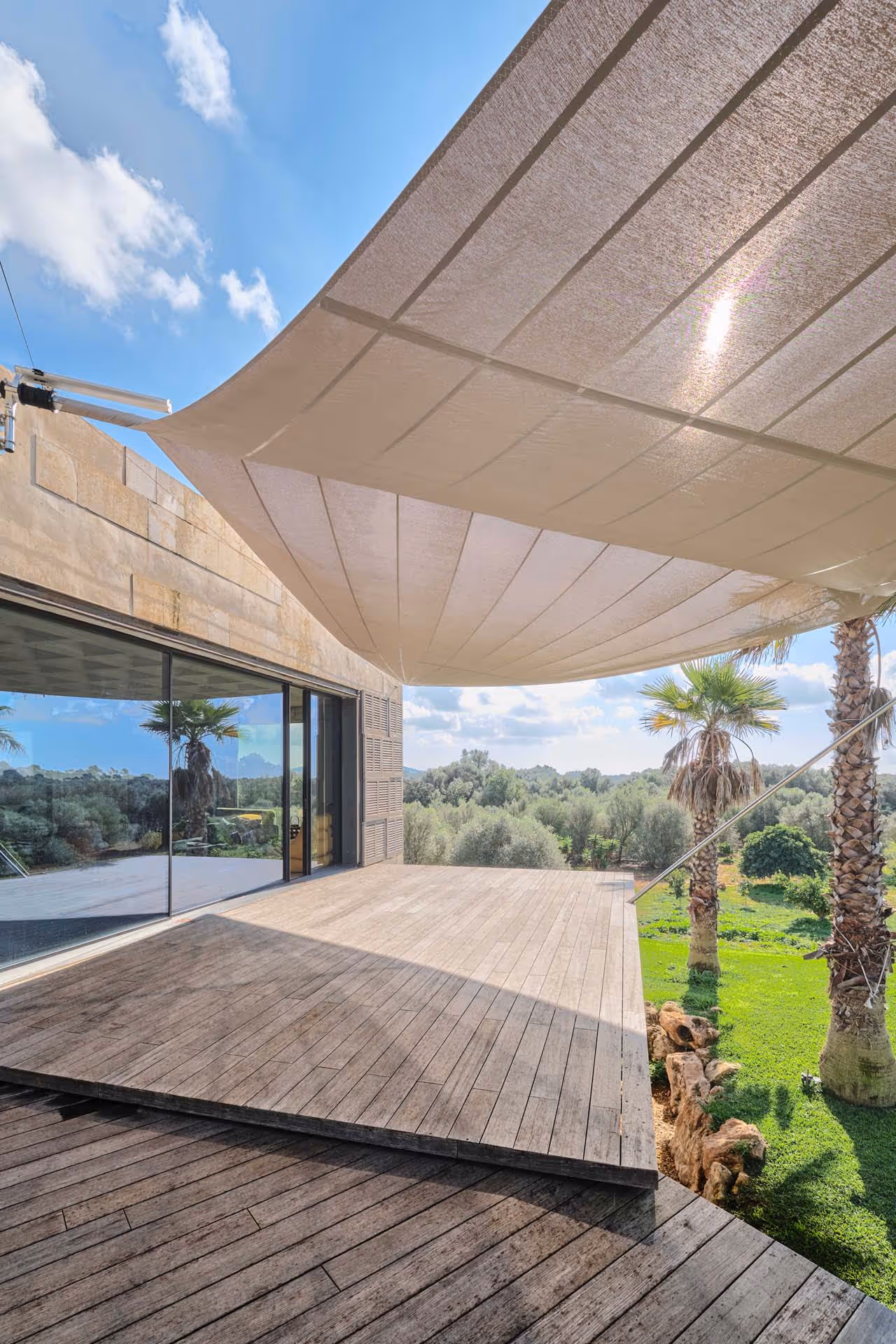 Stylish villa with sandstone façade and floating wooden deck, SunSquare awning over terrace and infinity pool with mosaic tiles, surrounded by a tropical garden with palm trees and Mediterranean plants.