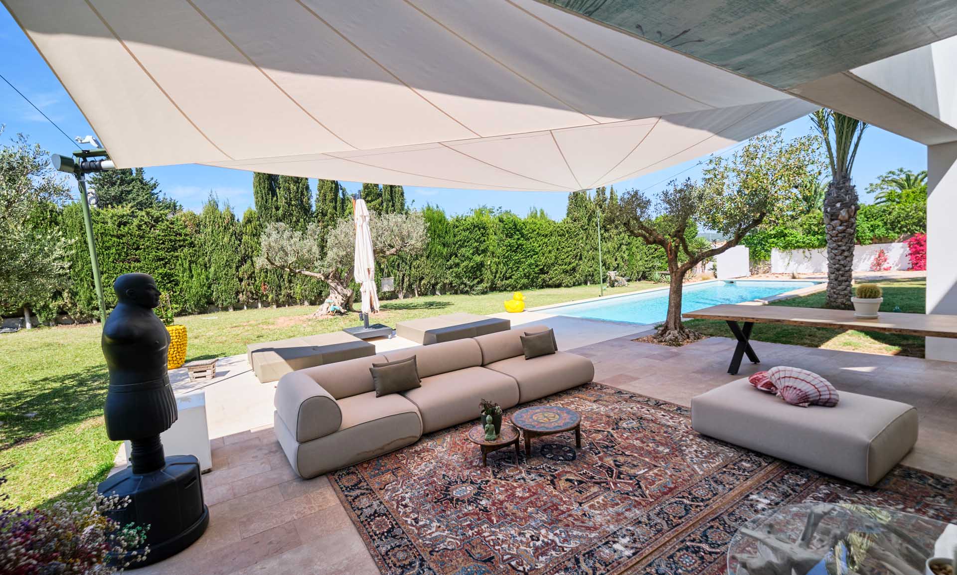 A stylishly designed outdoor area with olive trees, outdoor sofa and carpet harmoniously protected from sun and rain by a SunSquare shade sail.
