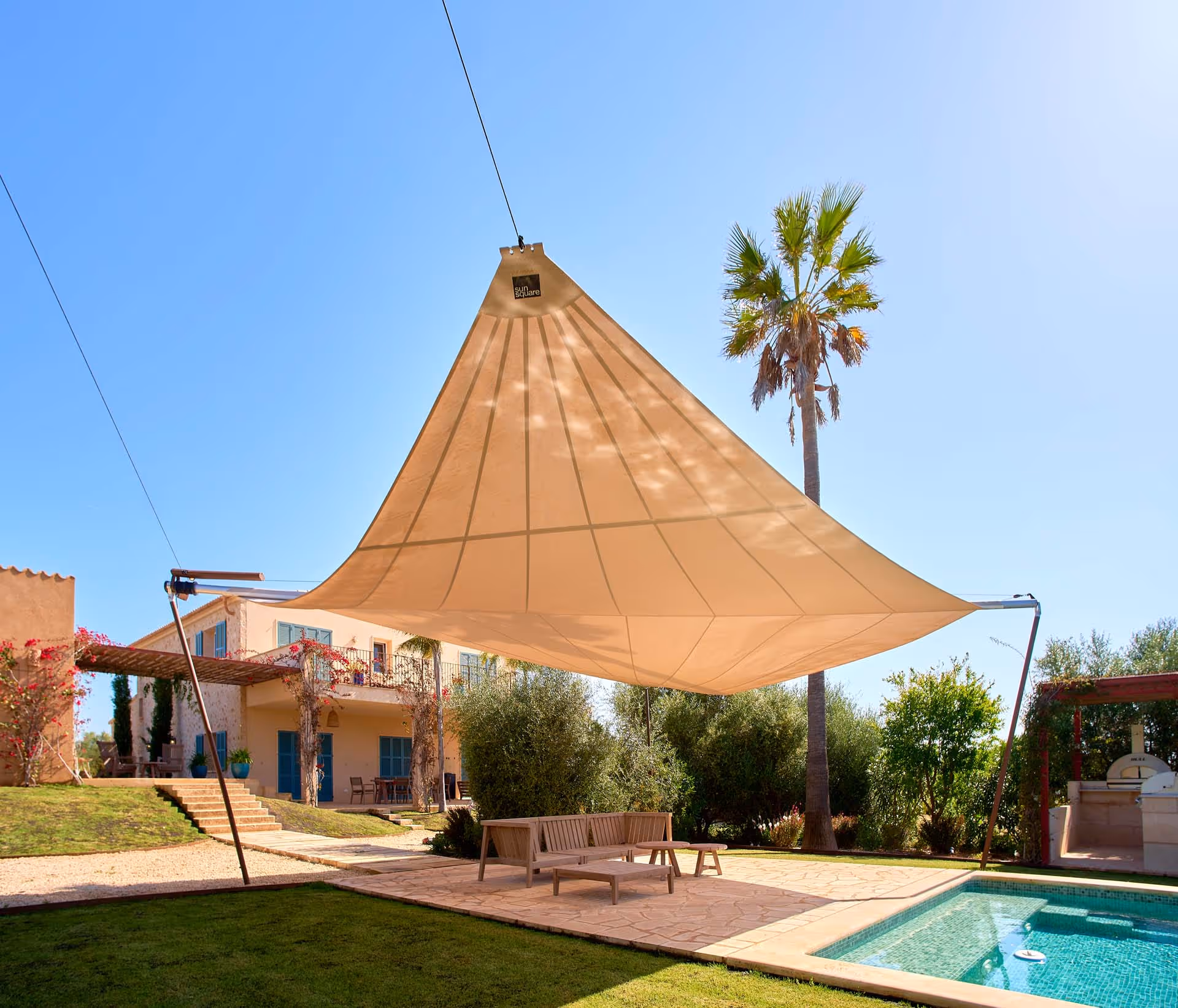 Powder-coated SunSquare awning system with four ground supports and radially cut sail, installed over a garden and pool area.