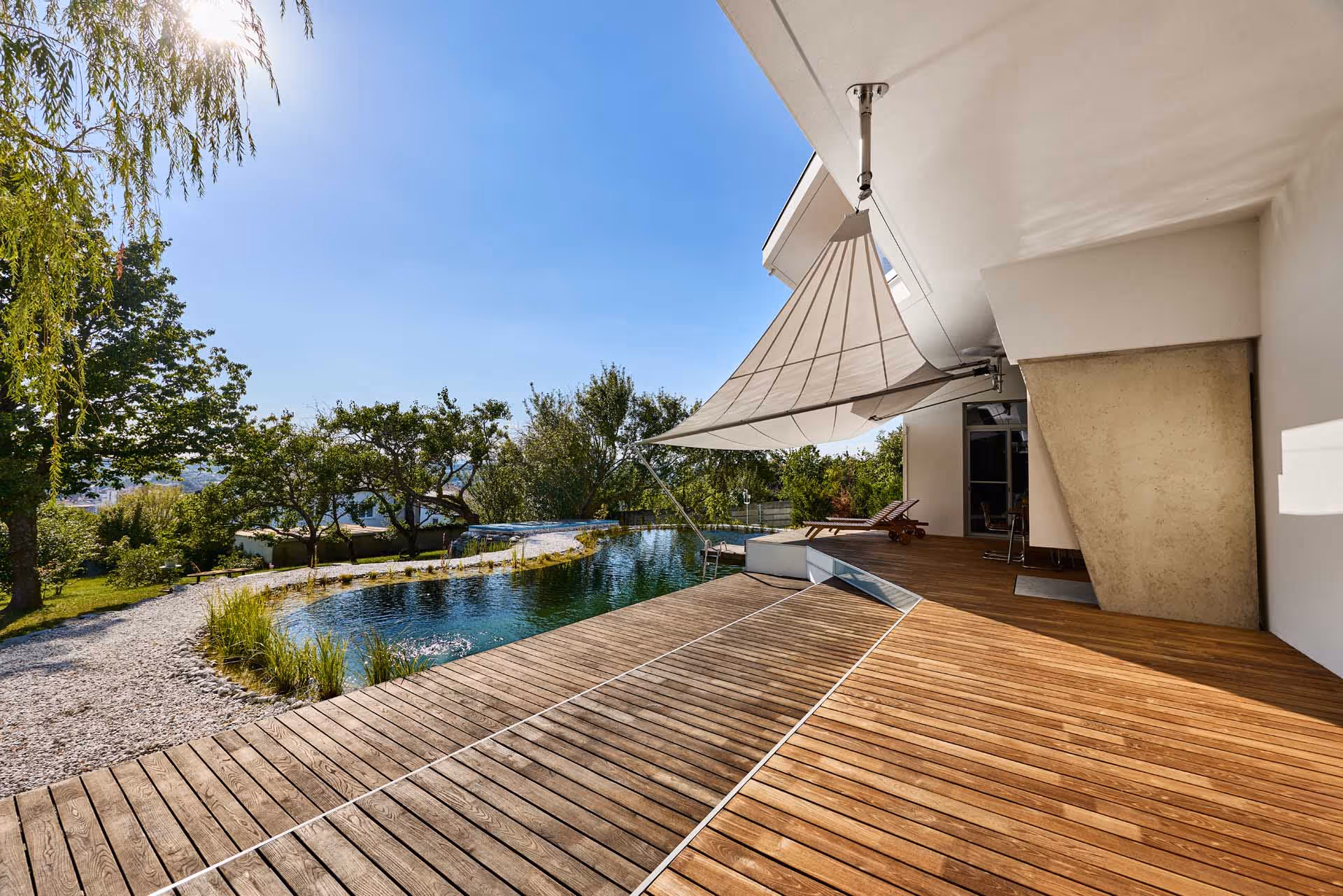 Sunny wooden deck terrace in two levels with adjacent swimming pond and SunSquare awning to protect the afternoon sun from overheating and the terrace and interiors.