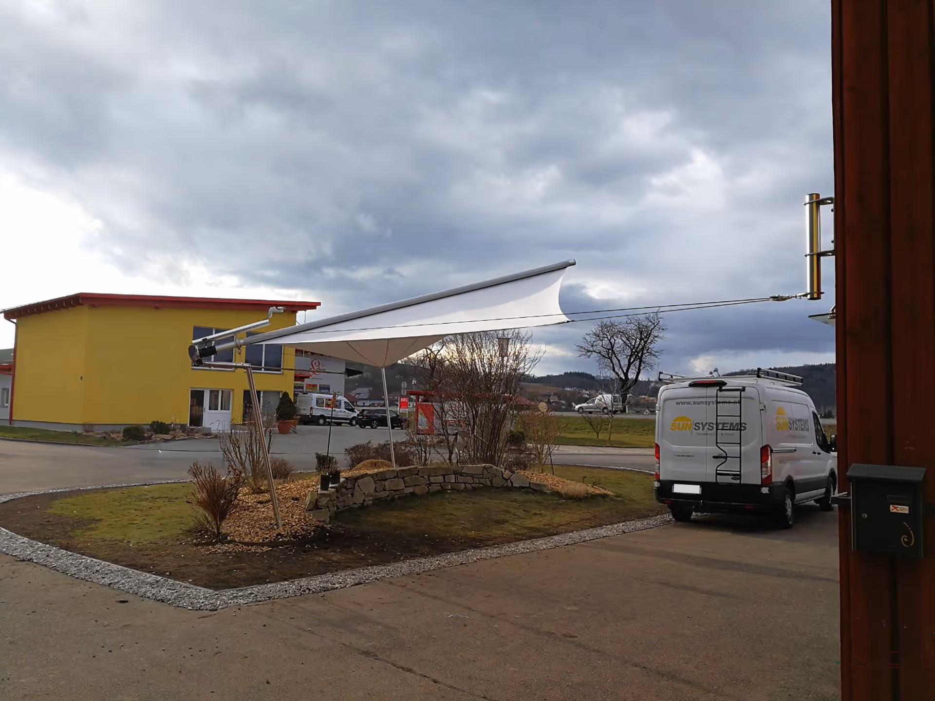 Freikragendes SunSquare Dreiceksegel mit 28 m² Fläche fügt sich elegant in den offenen Platz ein.