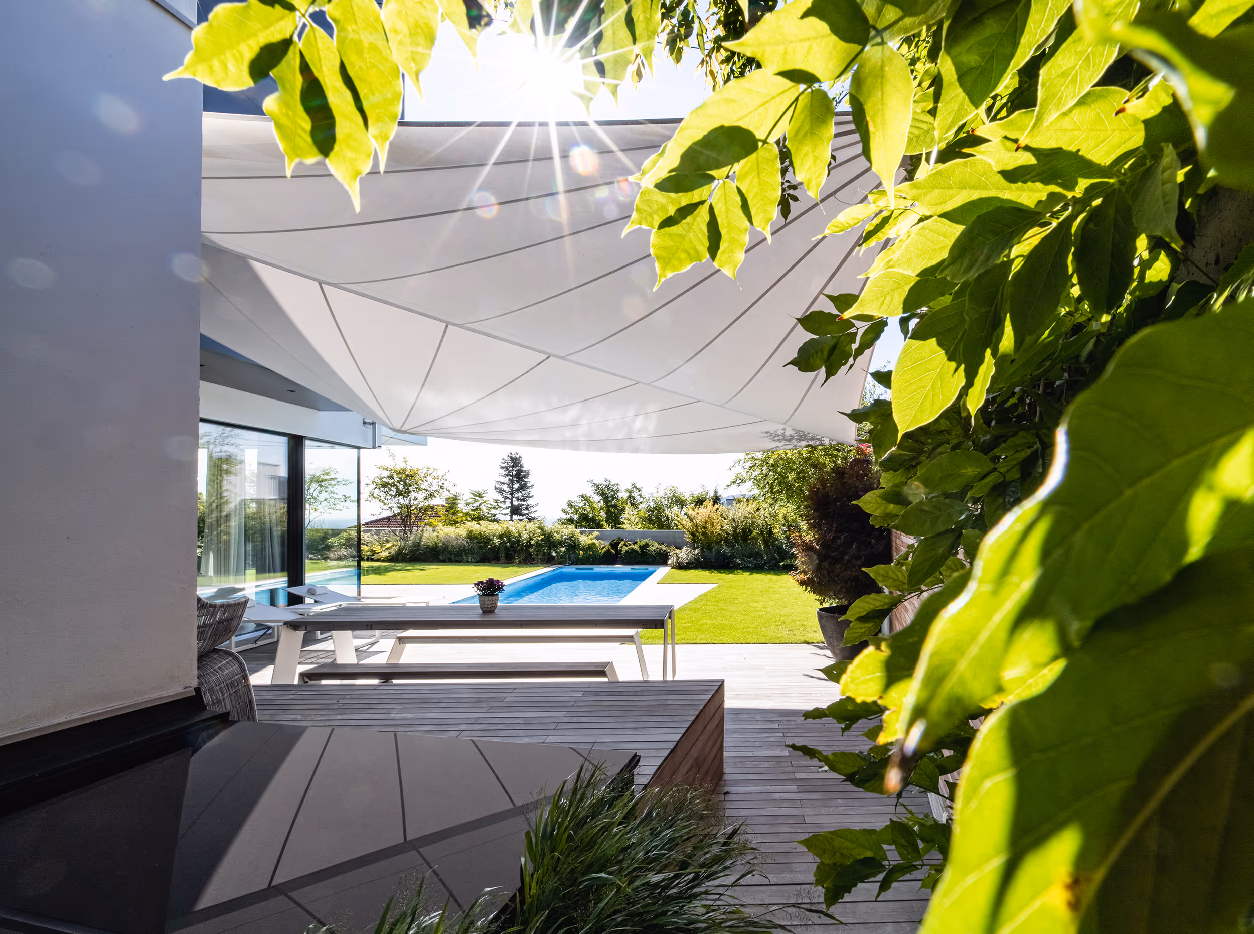 A tastefully designed garden with wooden deck, dining area, and adjacent swimming pool is protected from sun and rain by a SunSquare sail.