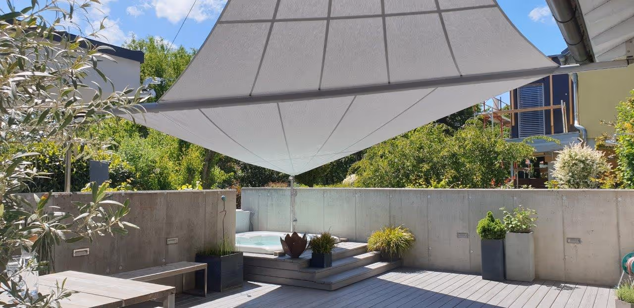 Spacious outdoor area with 24 m² awning, mounted with minimal supports over a wooden deck with integrated whirlpool. Surrounded by concrete walls, plants and outdoor furniture.