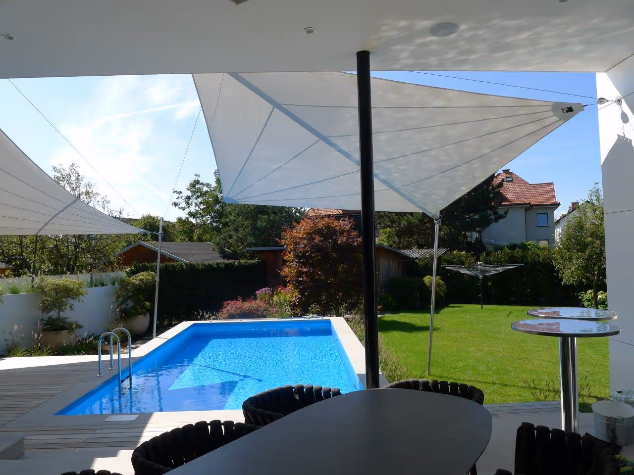 Ansprechender Außenbereich mit großen Sonnensegeln, die eine Holzterrasse und einen Pool beschatten – umgeben von üppigem Grün.
