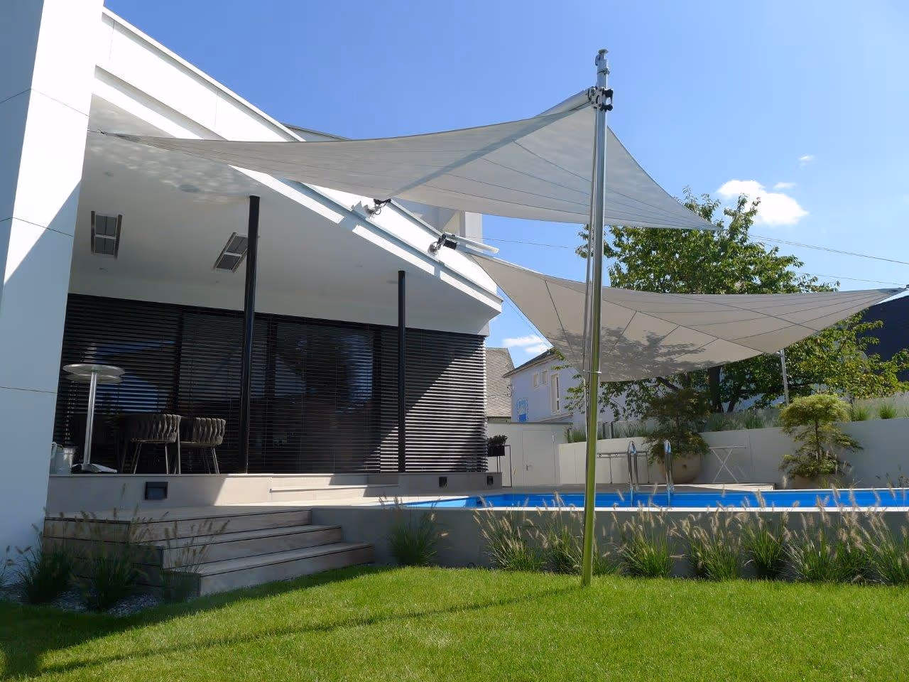 Moderner Außenbereich mit großen Sonnensegeln, die eine Holzterrasse und einen Pool beschatten – umgeben von üppigem Grün.