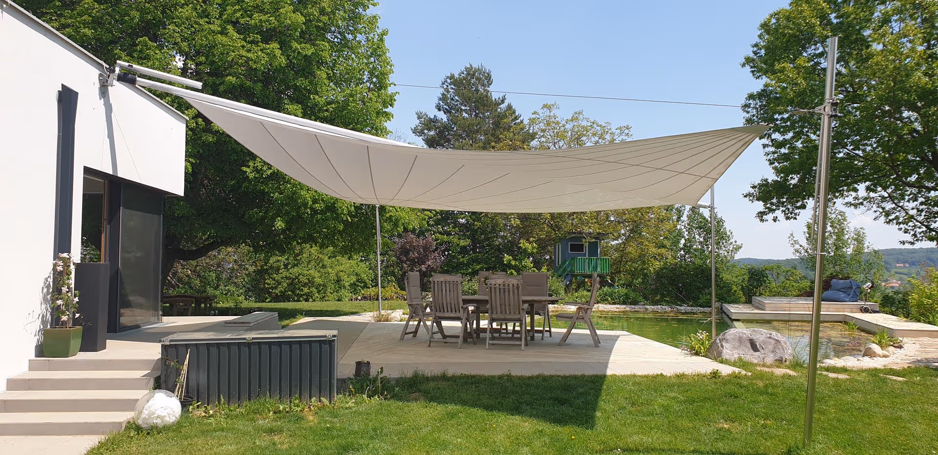 Man sieht aus einer seitlichen Perspektive eine große Glasfront des Hauses und die Montagepunkte des Sonnensegels und die beeindruckende Größe der Segelfläche.