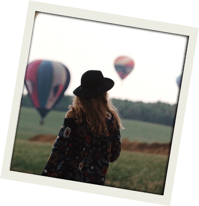 Woman with long hair and a black hat stands in a grassy field watching colorful hot air balloons rise into the sky. Framed like a vintage photo, capturing a serene travel or festival experience.