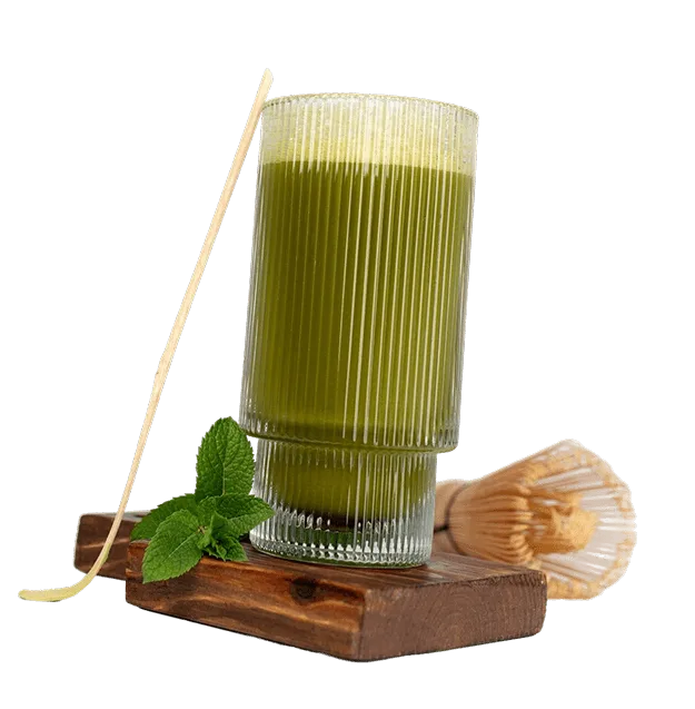 A tall ribbed glass filled with frothy green matcha tea, placed on a wooden board with a bamboo whisk, mint leaves, and a chasen stick. Aesthetic, healthy beverage setup.