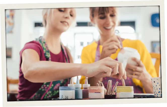 Two women smiling and painting ceramics at a pottery studio, surrounded by colorful paint jars and brushes. Fun, hands-on crafting experience.