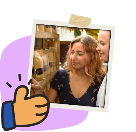 Two women smiling and examining packaged products on a shelf in a cozy store. Framed with a purple shape and a thumbs-up icon. Positive, community-focused shopping experience.