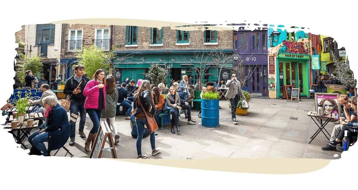 A lively courtyard filled with people sitting, chatting, and enjoying drinks outside colorful shops and cafes. Features eclectic storefronts, street art, and urban greenery. Social, creative community vibe.