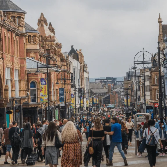 Busy urban street filled with pedestrians and shoppers, framed by ornate historic buildings and modern shop signs. Street lamps line the road under a cloudy sky. Vibrant city life and retail atmosphere.