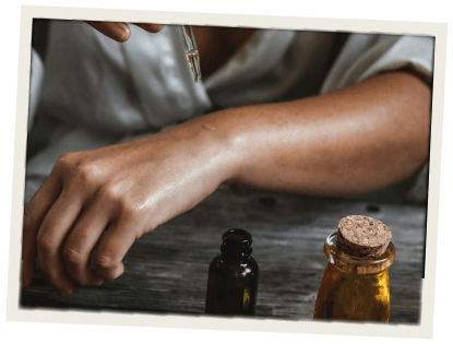 A close-up of a person applying essential oil to their arm using a dropper, with amber glass bottles nearby on a wooden surface.