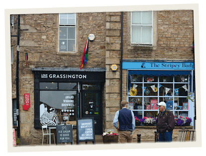 A charming village street with small independent shops, including “Love Grassington” and “The Stripey Badger,” with two men chatting outside.