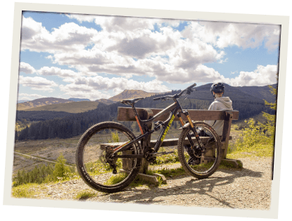 A scenic viewpoint with a mountain bike parked beside a bench and a cyclist admiring the landscape of hills and forests.