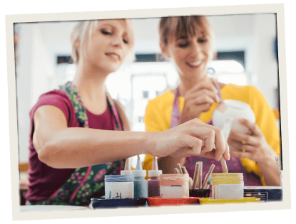 Two women in aprons smiling and painting pottery at a craft workshop table with colorful paints.