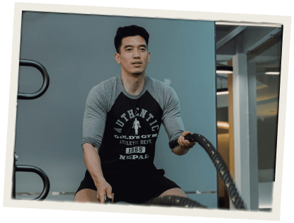 A man in a gym performing battle rope exercises, dressed in a workout shirt and shorts.