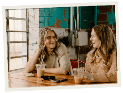 Two women enjoying iced coffee at a cozy, urban-style café with exposed brick and industrial decor.