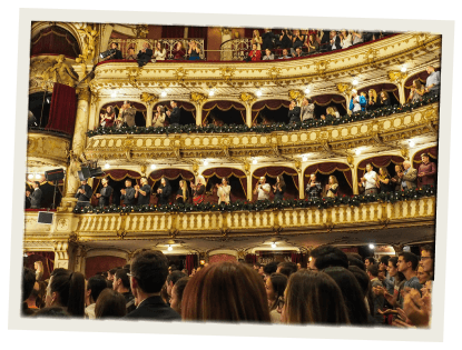 A grand, ornate theatre filled with people clapping and standing during a performance or event.