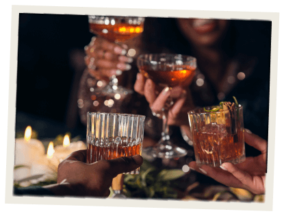 A lively group toast featuring a mix of elegant cocktail glasses filled with amber-colored drinks. The mood is festive and warm, with candlelight and smiling faces in the background.