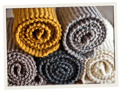 A neatly stacked display of rolled-up, chunky knitted blankets in earthy tones like mustard, grey, brown, and cream. Perfect for a cozy, handcrafted product showcase.