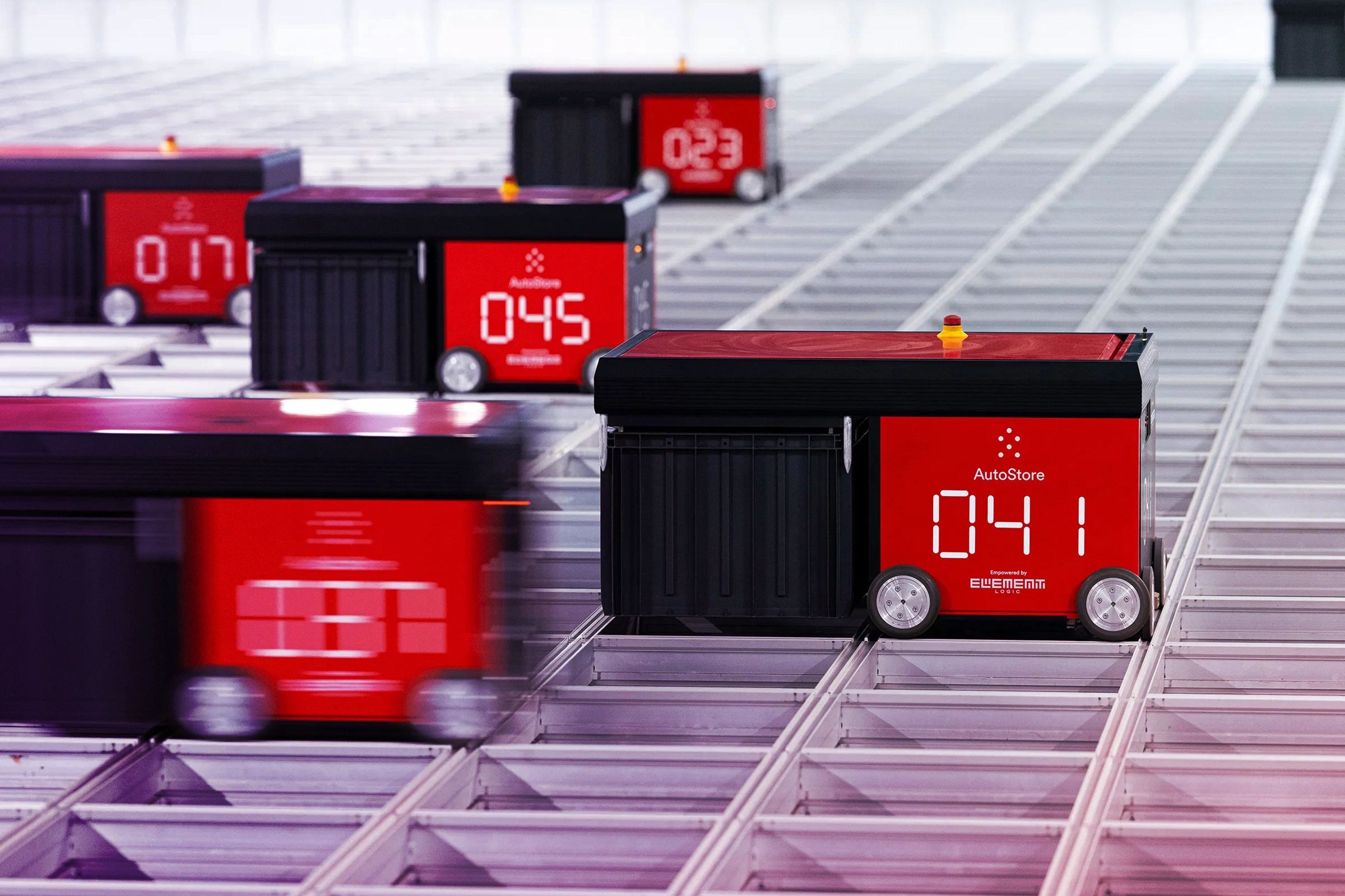 Several red and black AutoStore robots moving along a grid system in a warehouse environment.