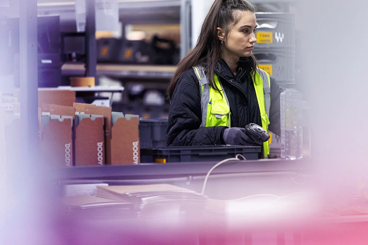 Woman wearing a neon safety vest and gloves holding a barcode scanner in a warehouse packed with boxes and plastic crates.