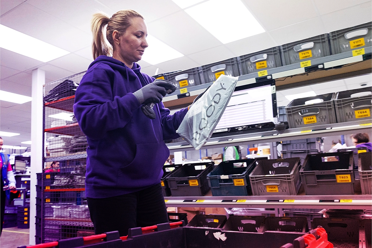 Woman in purple hoodie scanning a package labeled 'BCODY' in a warehouse with shelves of bins.