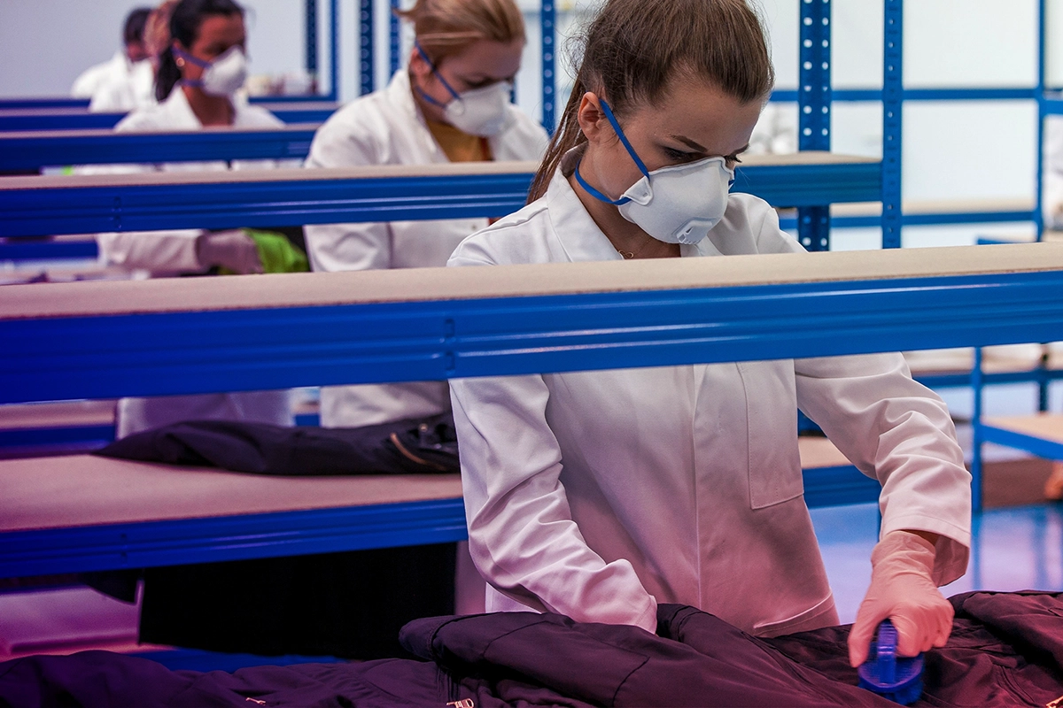 Workers wearing white lab coats and face masks cleaning or inspecting dark jackets on blue metal racks in a factory setting.