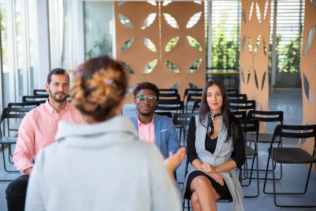 Group training session with a diverse audience engaged in sales and leadership development, featuring a woman presenting to attentive participants in a modern classroom setting.