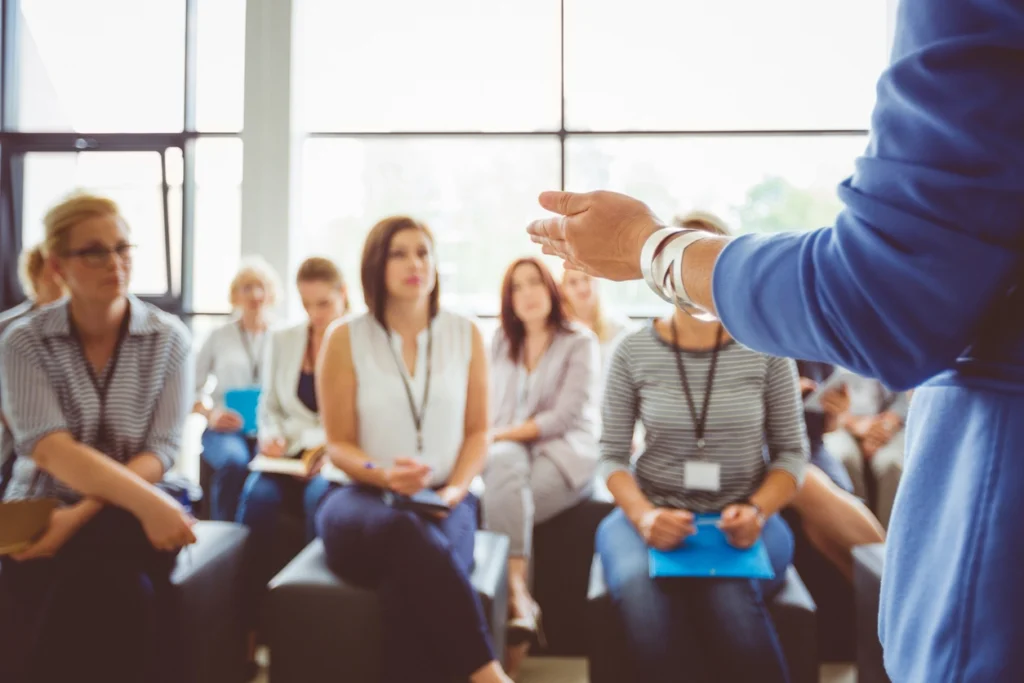 Group of diverse professionals engaged in a leadership training session, with a speaker gesturing, emphasizing the importance of effective leadership development.