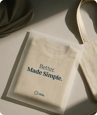 Folded beige t-shirt with the text 'Better. Made Simple.' and the Vela logo lying on a light surface next to a beige tote bag and fabric.