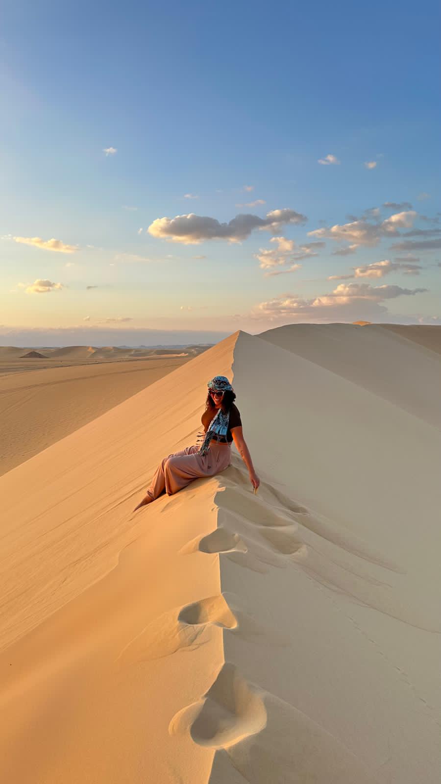  Frau sitzt entspannt bei Sonnenuntergang auf einer goldenen Sanddüne in der Weißen Wüste Ägyptens – ein stiller Moment inmitten der atemberaubenden Wüstenlandschaft.
