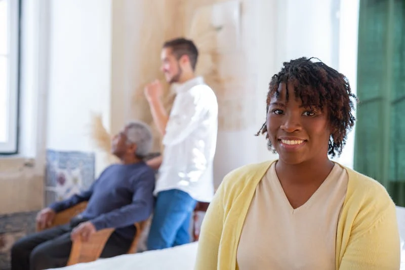 A woman smiles confidently while a caregiver tends to an elderly man in the background.