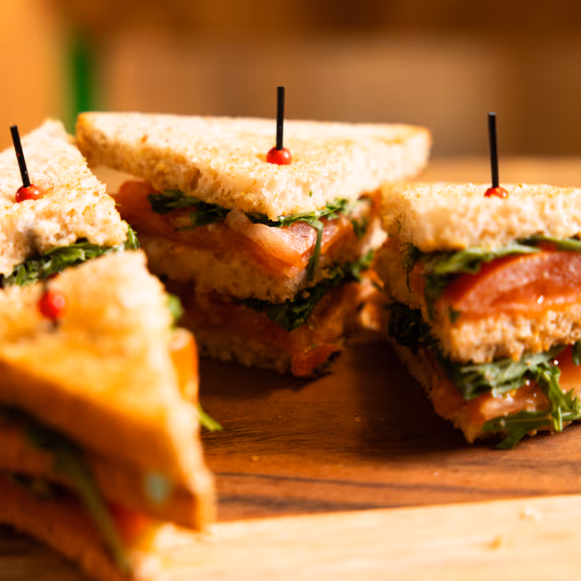 Sandwichs en triangles garnis de laitue et tomate tenus par des pics rouges sur planche en bois.