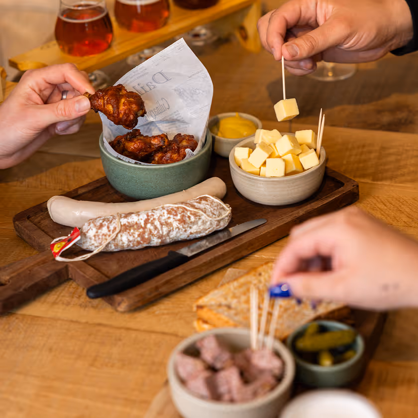 Plateau avec saucisses, cubes de fromage, ailes de poulet, moutarde et cornichons, mains prennent de la nourriture.