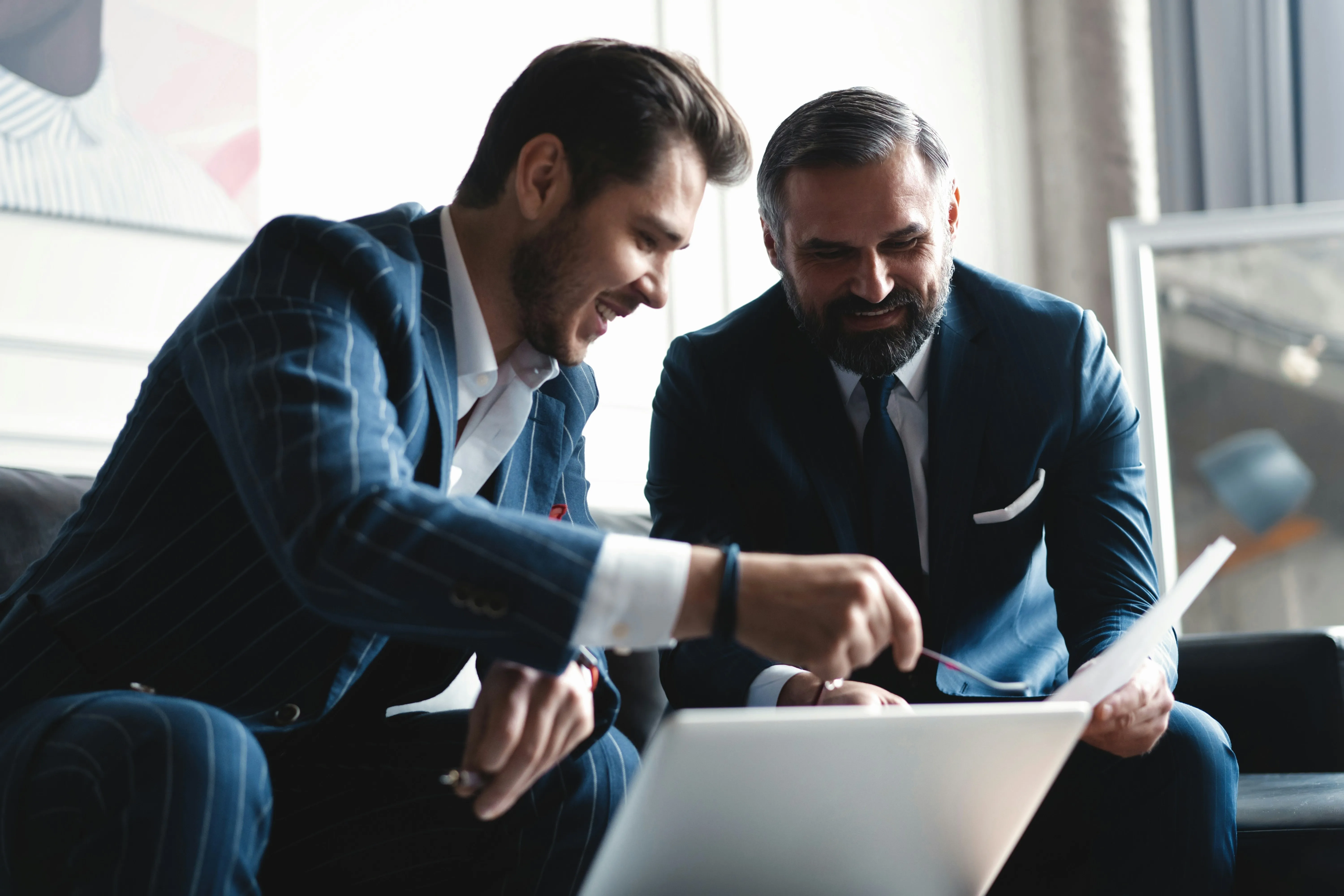 Two men in suits smiling and discussing documents while looking at a laptop in an office.