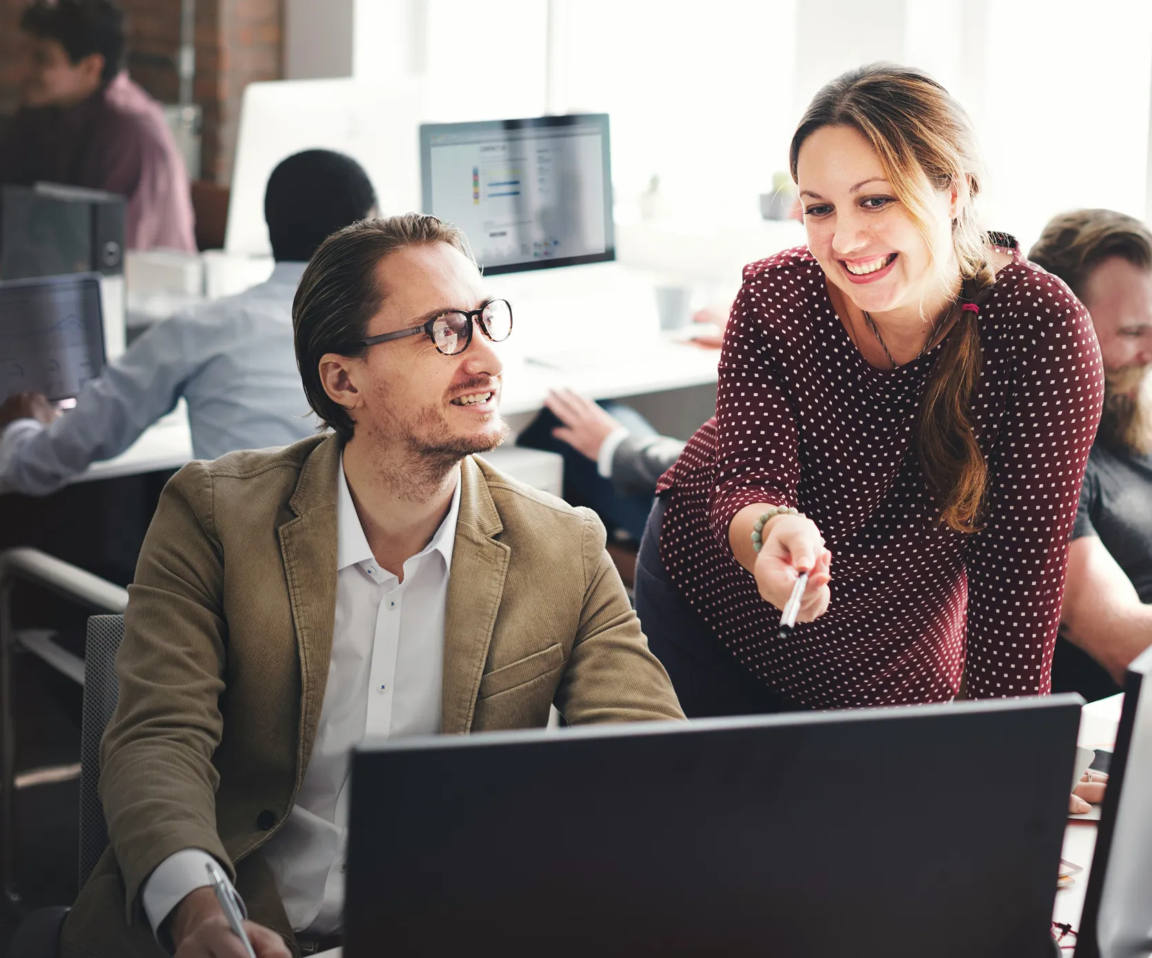 A woman in a maroon dotted blouse points at a computer screen while smiling and talking to a seated man wearing glasses and a brown blazer in an office.