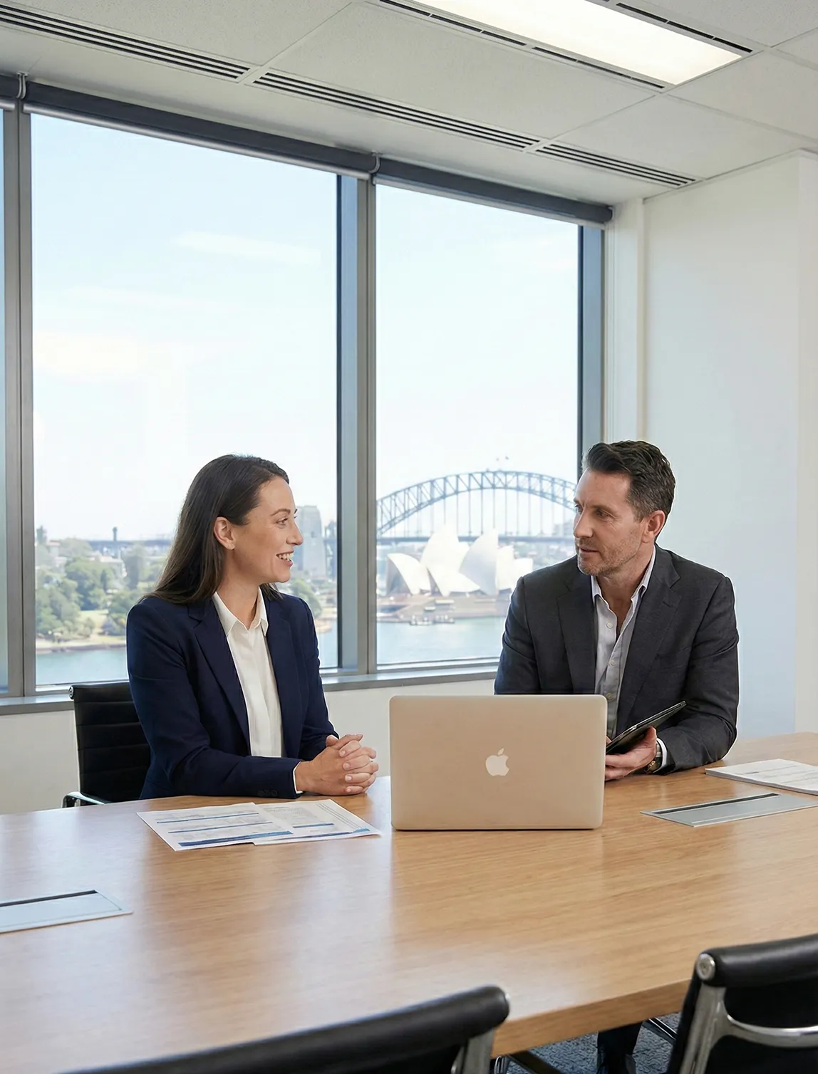 Two business professionals in suits having a discussion at a conference table with a laptop, Sydney Opera House and Harbour Bridge visible through the window behind them.
