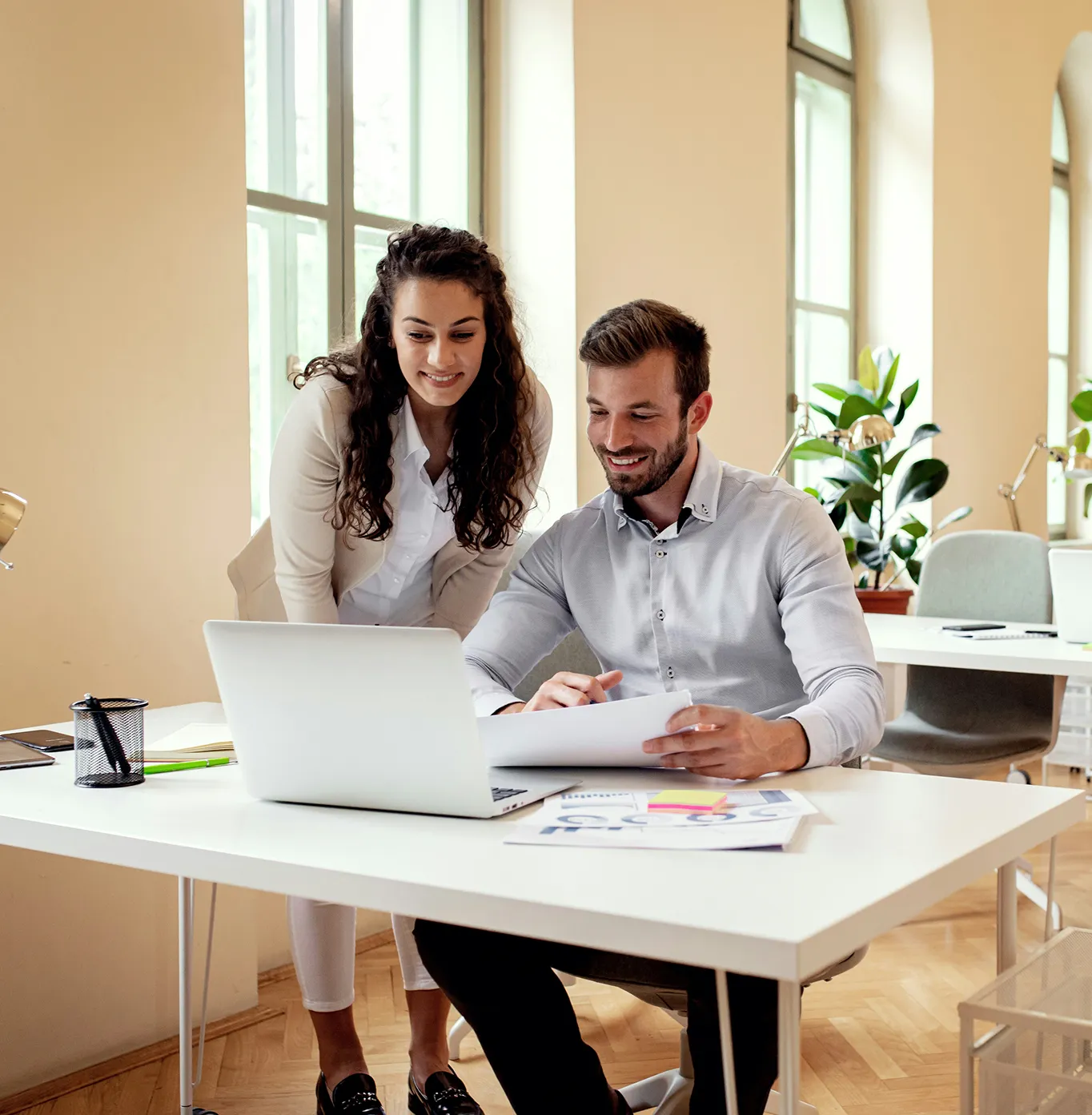 Businessman and businesswoman reviewing documents together at a desk with a laptop in a bright office.