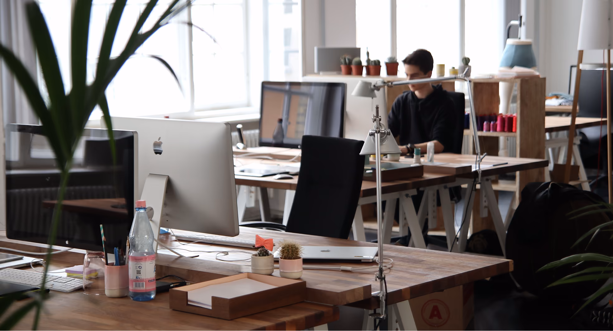 Modern office with wooden desks, computers, small plants, and a person working in the background.