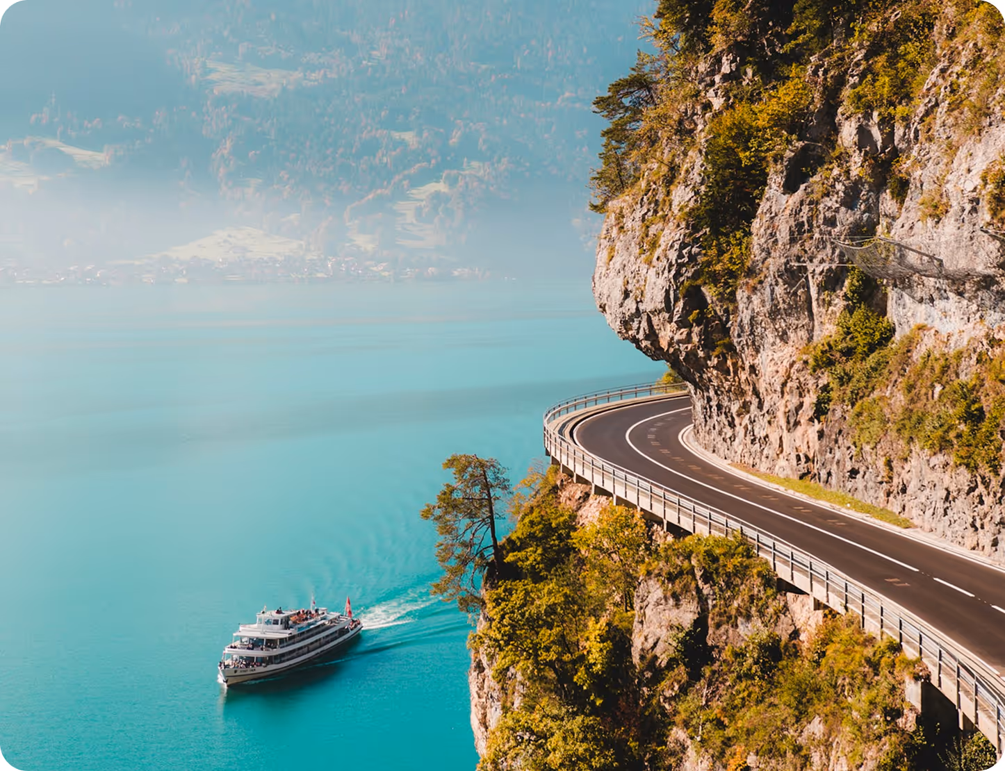 Tour boat sailing on turquoise lake near a winding road on a rocky cliff with green trees.