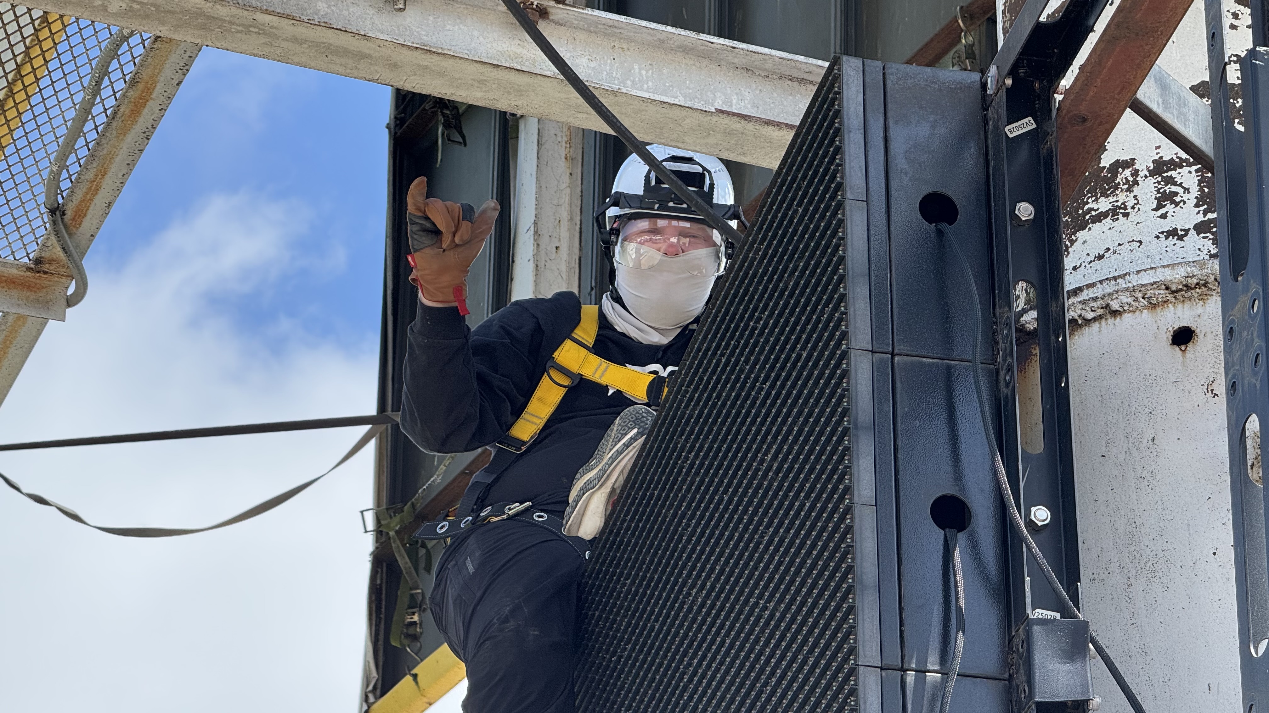 A worker wearing safety gear, including a helmet, face covering, gloves, and a yellow harness, gives a hand signal while removing an LED screen panel on a metal structure outdoors.
