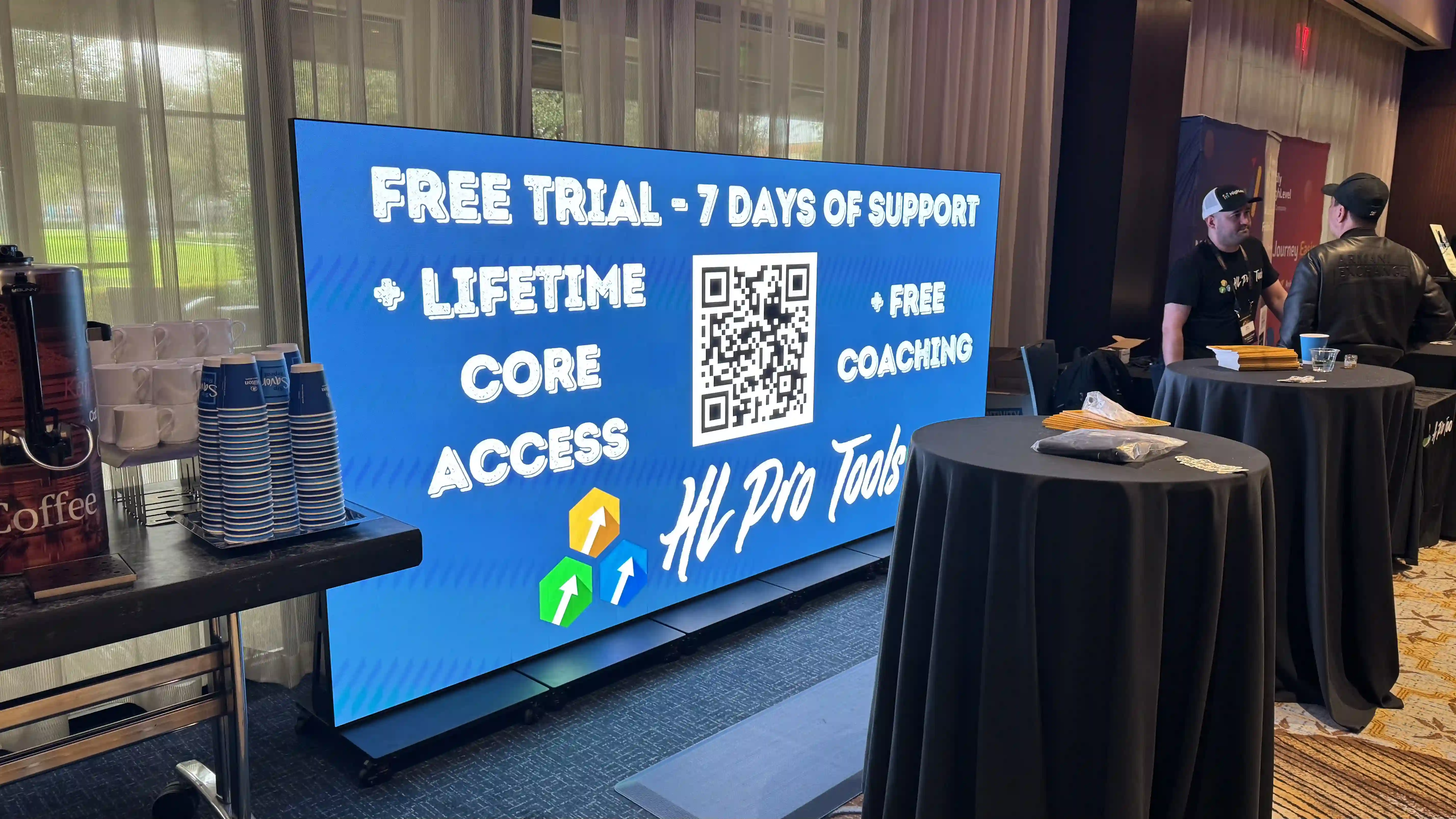 Portable LED display screen at a trade show booth showing a promotional message and QR code, surrounded by coffee, branded cups, and attendees conversing nearby.