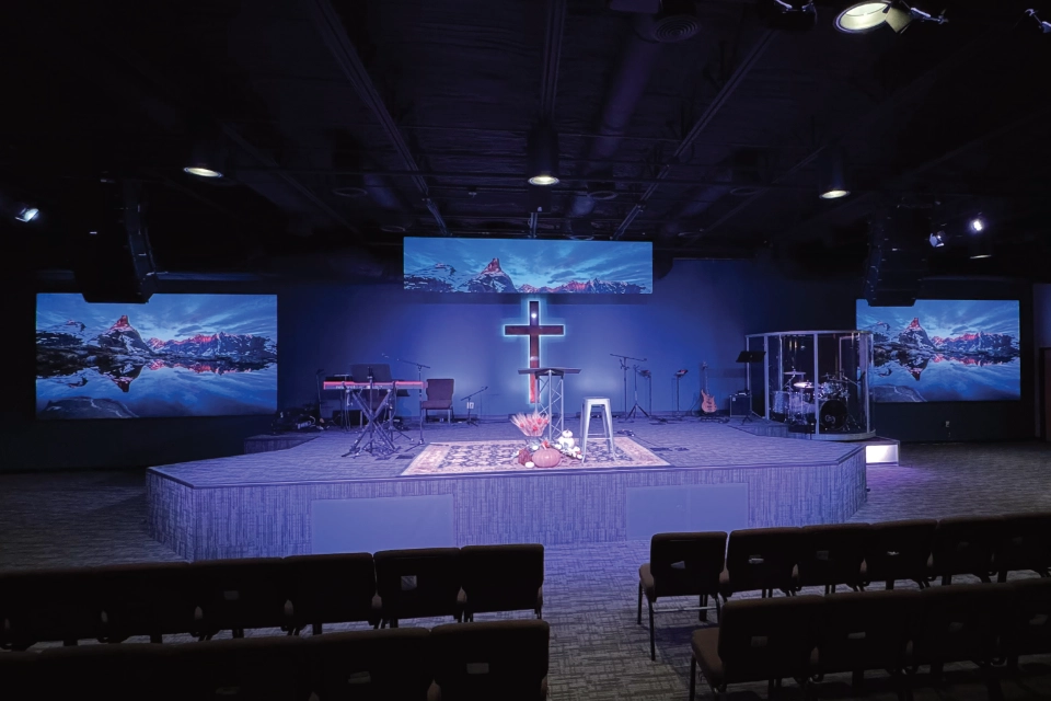 Modern church stage setup with multiple LED screens displaying a scenic mountain image, surrounding a central cross and worship instruments.