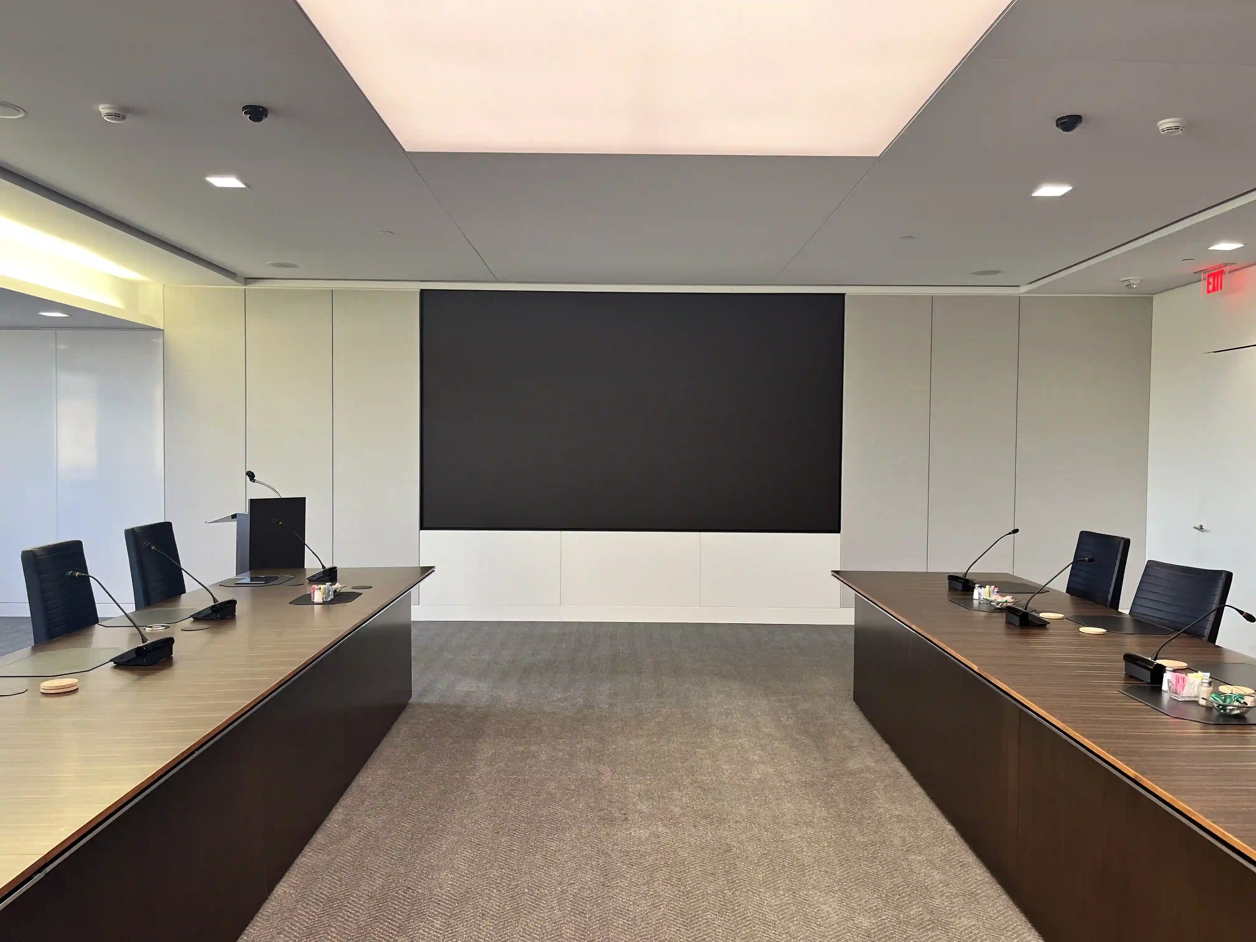Minimalist corporate boardroom with a large black LED display centered on the wall between two sleek wooden conference tables with microphones.