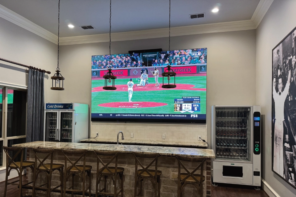 Residential bar area with a mounted LED screen showing a baseball game, surrounded by vending machines, a drink cooler, and seating.