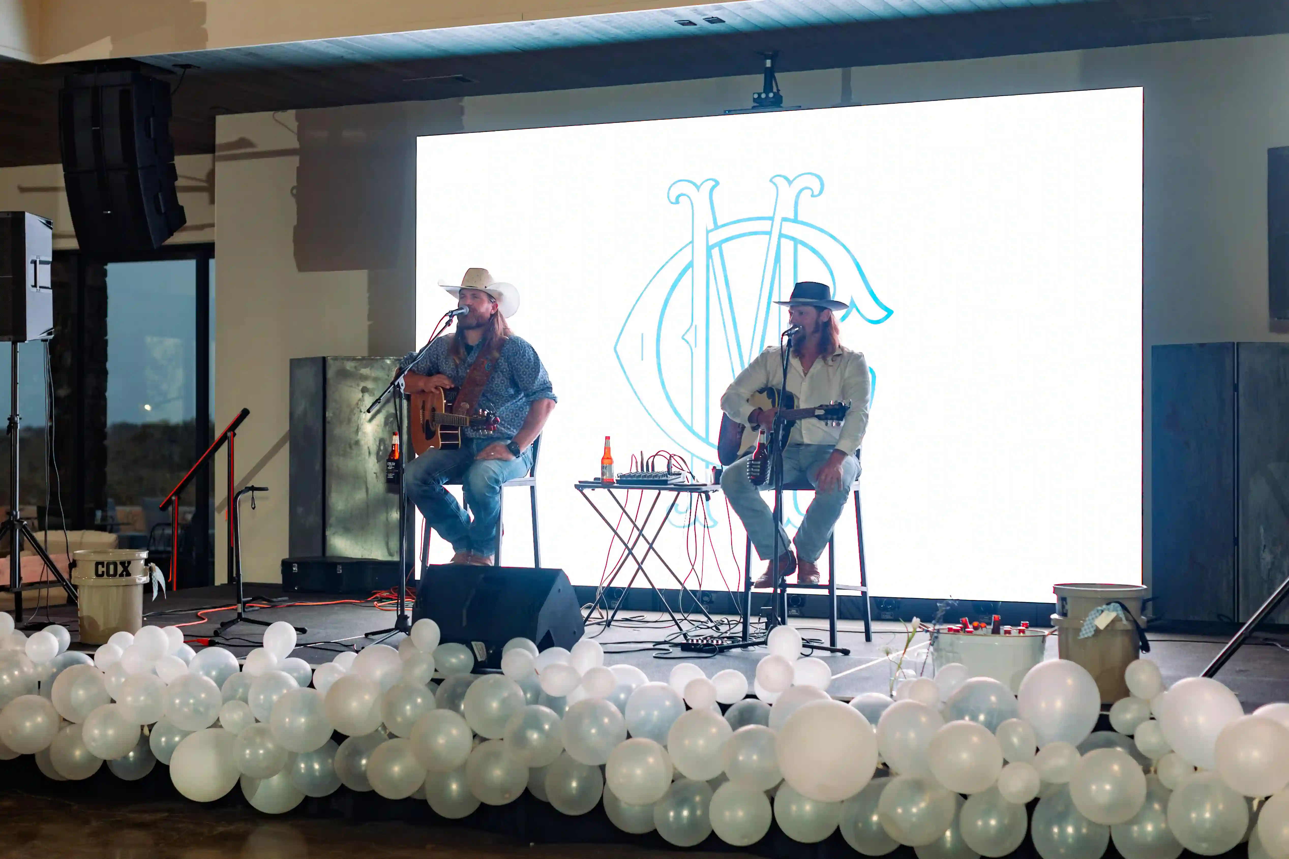 Two musicians performing on stage with acoustic guitars in front of a bright LED video wall displaying a monogram logo, surrounded by white balloons at an indoor event venue.